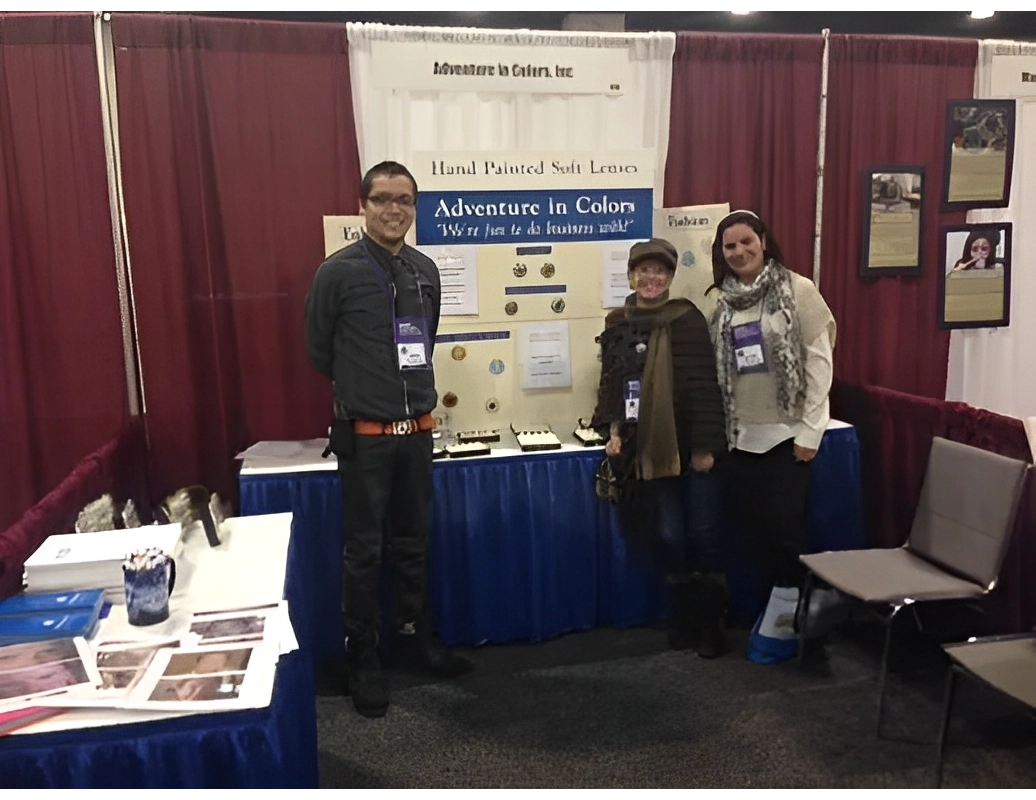 Three people standing in front of a booth display with the sign "Adventure in Colors, Inc." in a convention setting.