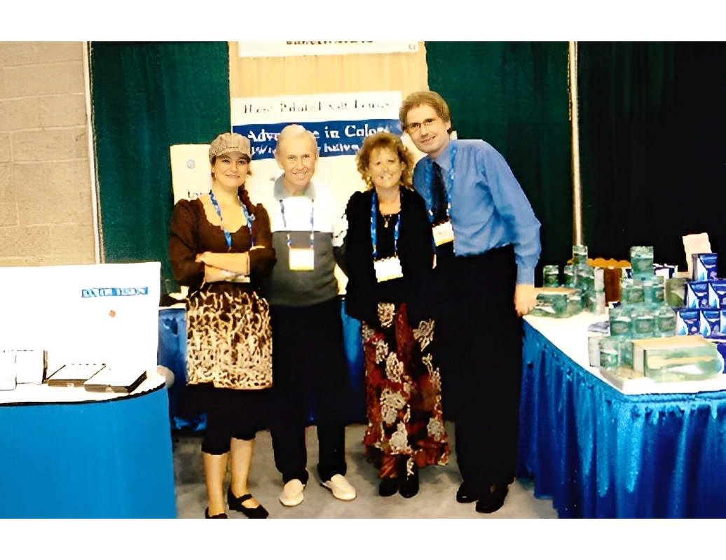 Four people stand together in a trade show booth, smiling at the camera. The booth features banners, brochures, and product displays on a table with a blue tablecloth.