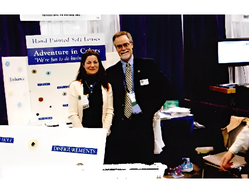 Two people stand in a booth at a convention with signage about hand-painted soft lenses. Some displays and chairs are visible nearby.