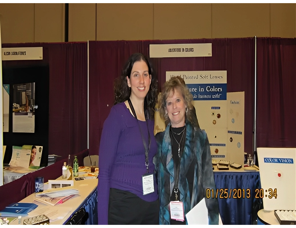 Two women smiling in front of a booth at a convention, wearing name badges. Behind them are display boards with text and images.
