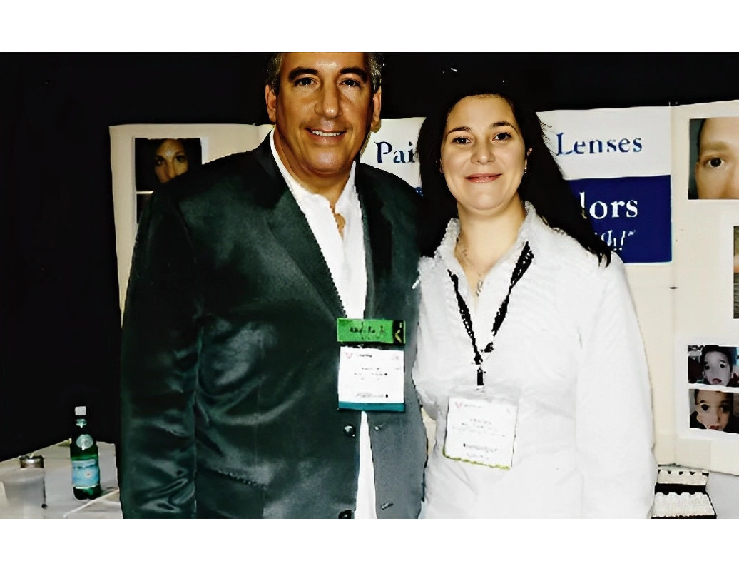 Two people smiling, wearing badges, standing in front of a booth with signs about lenses.
