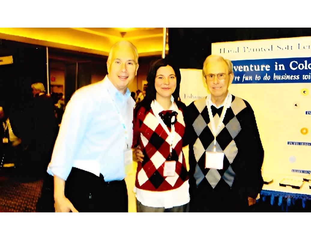 Three people stand together indoors, smiling, in front of a booth with text about conferences. They wear lanyards and argyle sweaters.