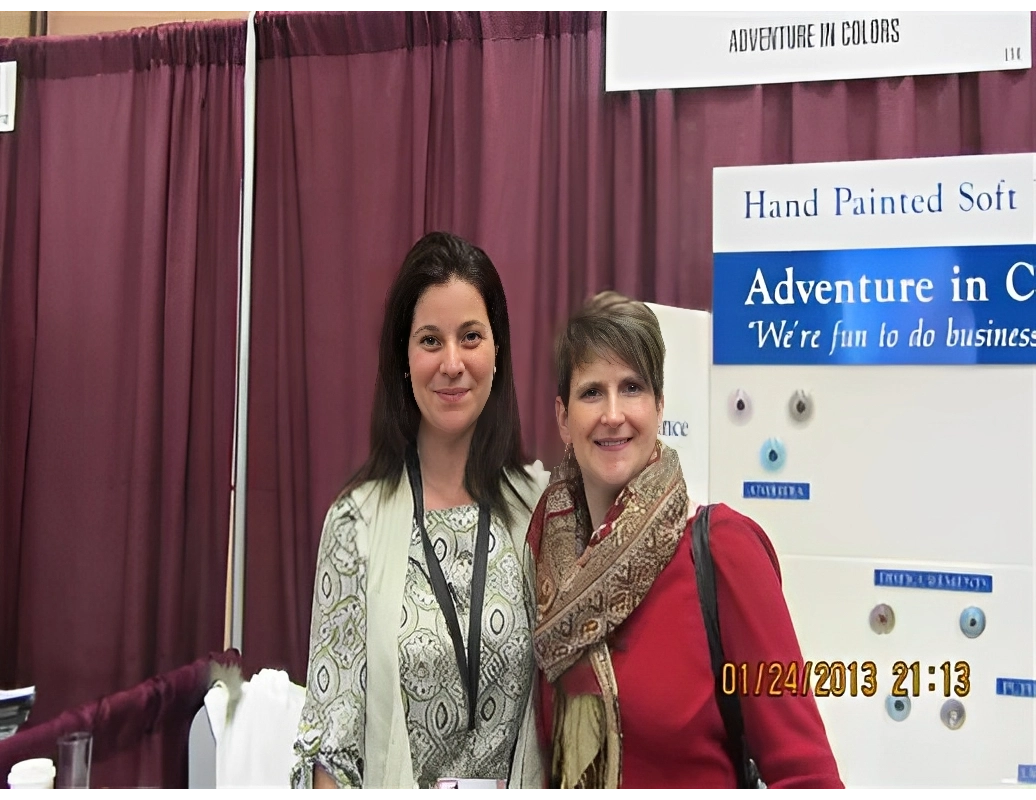 Two women stand smiling in front of a trade show booth displaying banners for "Adventure in Colors" and "Hand Painted Soft.