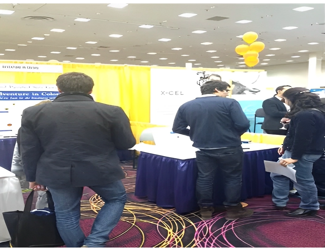 People at a convention booth with yellow curtains in the background. Balloons hang above. A person holds a paper, and another has a bag.