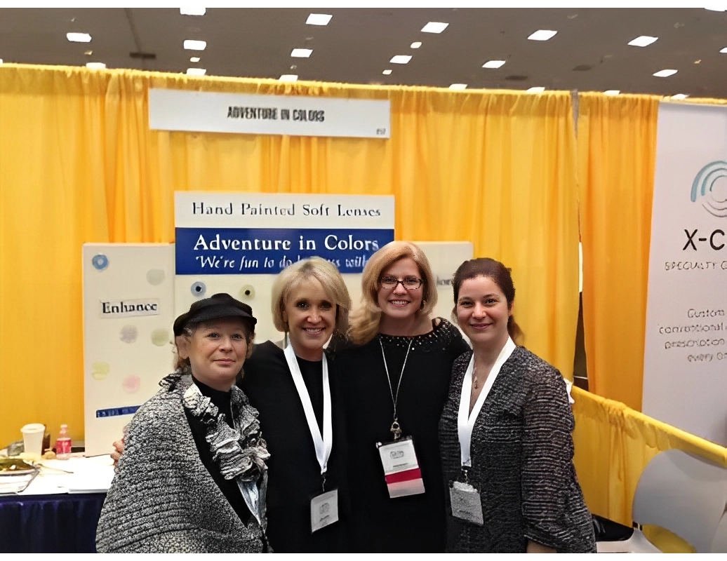 Four women smiling and standing together at a booth with a yellow backdrop, displaying "Adventure in Colors" and "Hand Painted Soft Lenses" signs.