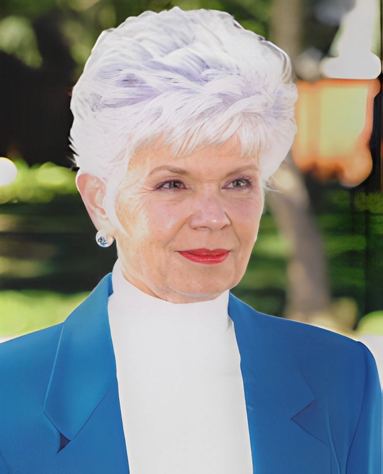 Elderly woman with short white hair, wearing a blue jacket and white turtleneck, standing outdoors with trees in the background.