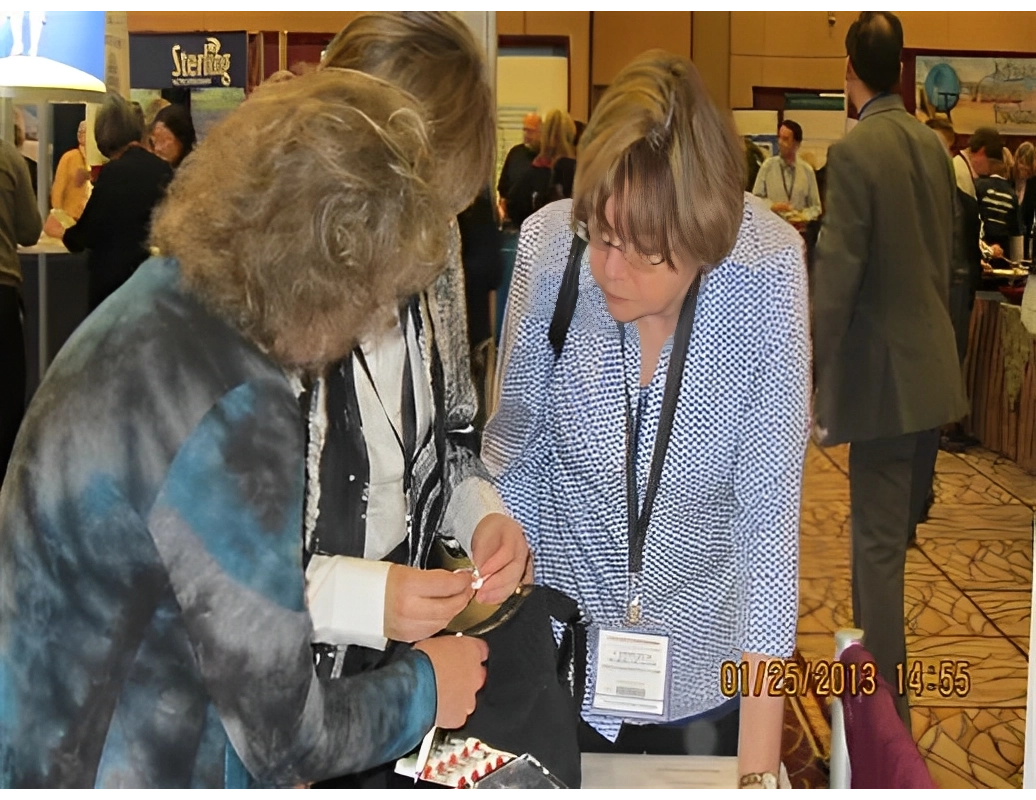 Three people closely examine an item at a conference. Two women hold badges, with one pointing at the object. People and exhibits are visible in the background. Date stamp: January 25, 2013, 14:55.
