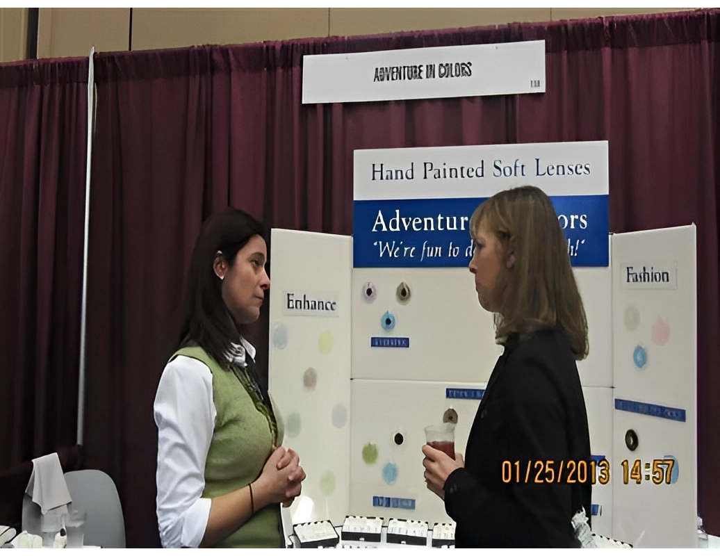 Two women are conversing in front of a booth with hand-painted soft lenses for enhancing eye color. The display includes text about enhancement, adventure, and fashion.