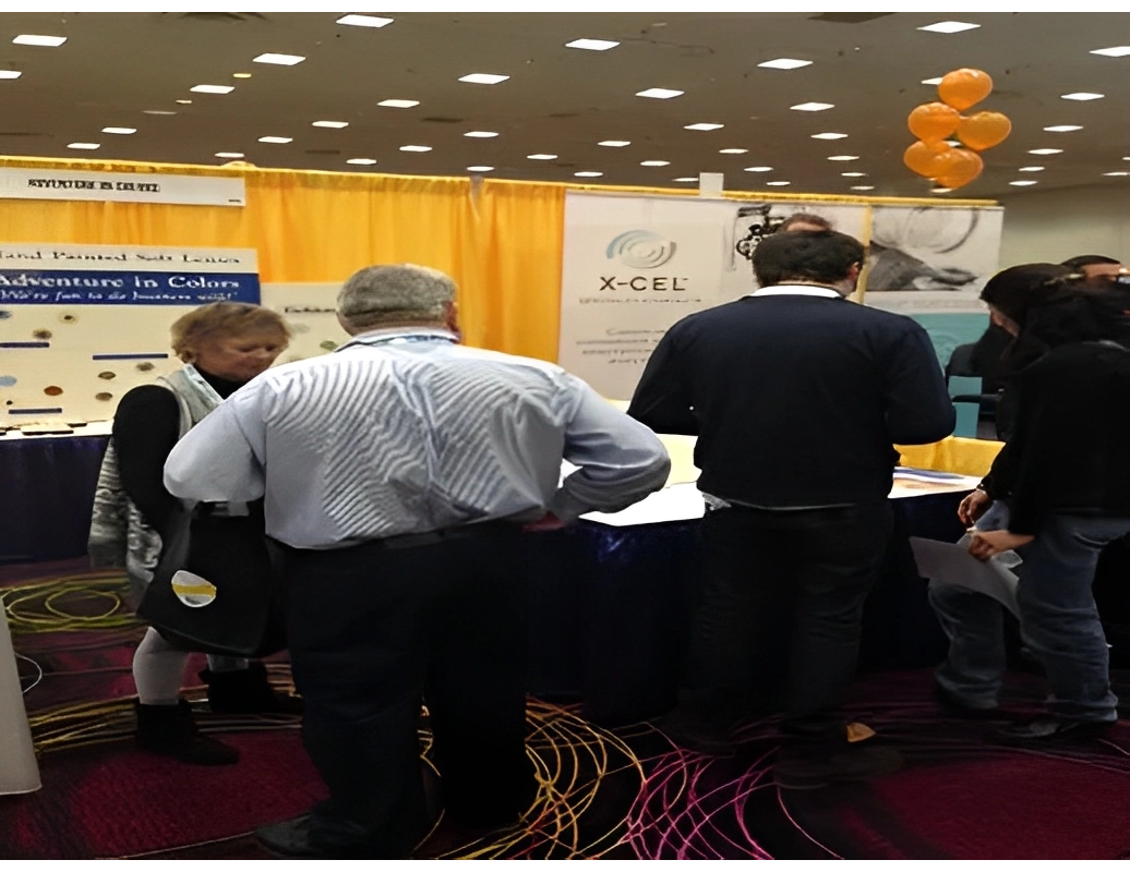 People gathered around a booth at a conference with informational displays and a cluster of orange balloons above.