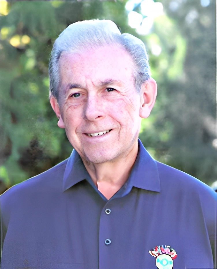 Elderly man with gray hair wearing a dark shirt with a crest, smiling outdoors with trees in the background.